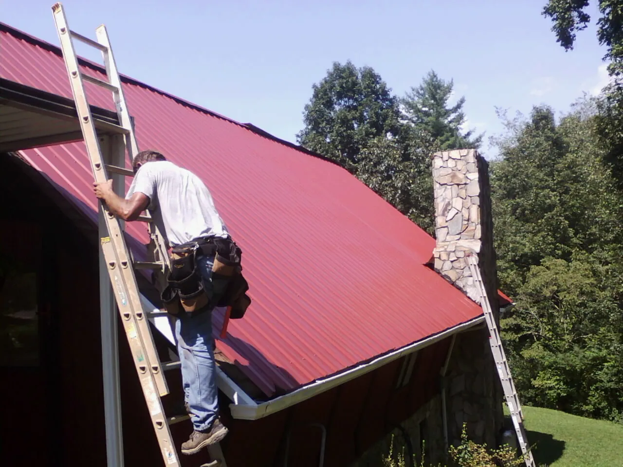 Roofer on a ladder beside a red metal roof and stone chimney — Soffit & Fascia Repair by Roofing Brothers in Quaker City